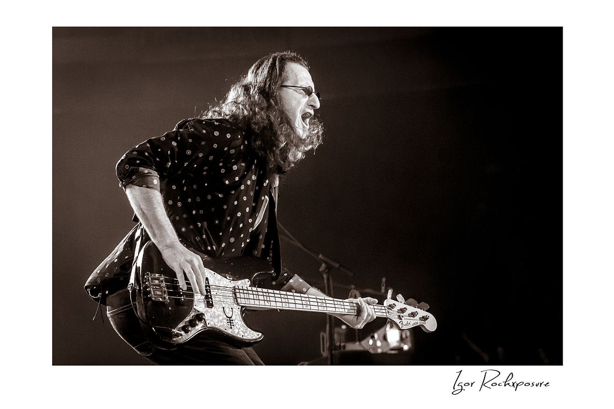 Horizontal concert photography of Geddy Lee leaning forward while playing bass guitar under stage lighting