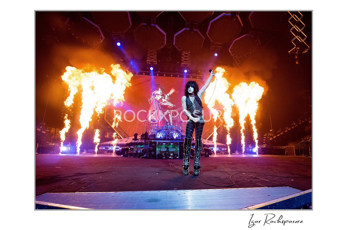 Paul Stanley of KISS standing with arm raised on stage while tall flames erupt behind him under dramatic red concert lighting