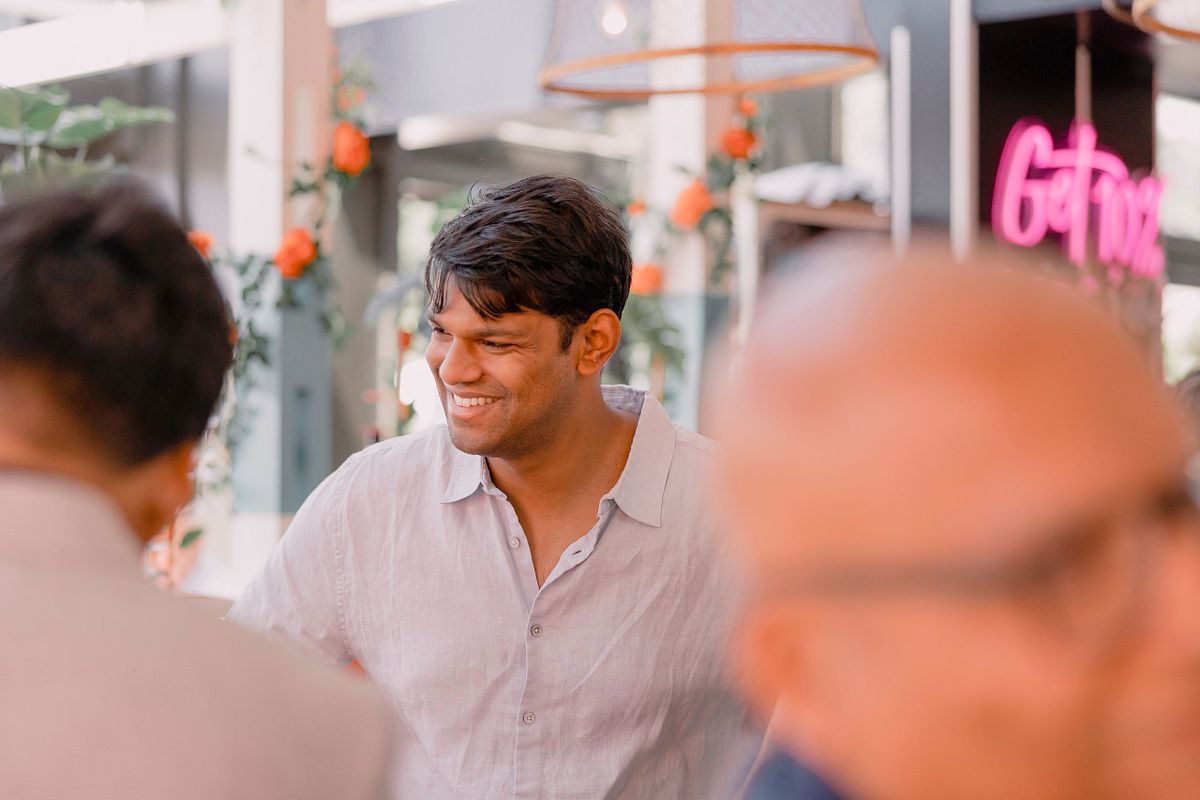 Photographer in Basel capturing a man at an engagement party in a light shirt smiles warmly in a lively, floral-decorated setting. Soft focus foreground figures add depth. Bright and cheerful atmosphere.