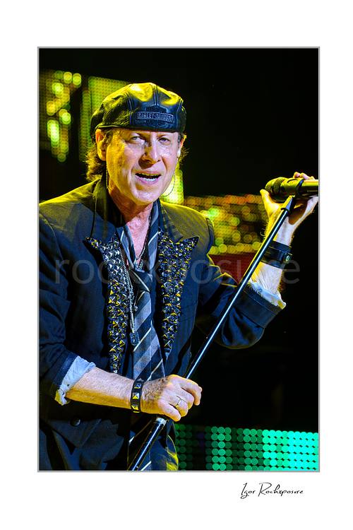 Vertical color image of Klaus Meine of Scorpions singing live with microphone stand, black cap, and sparkling green stage lights